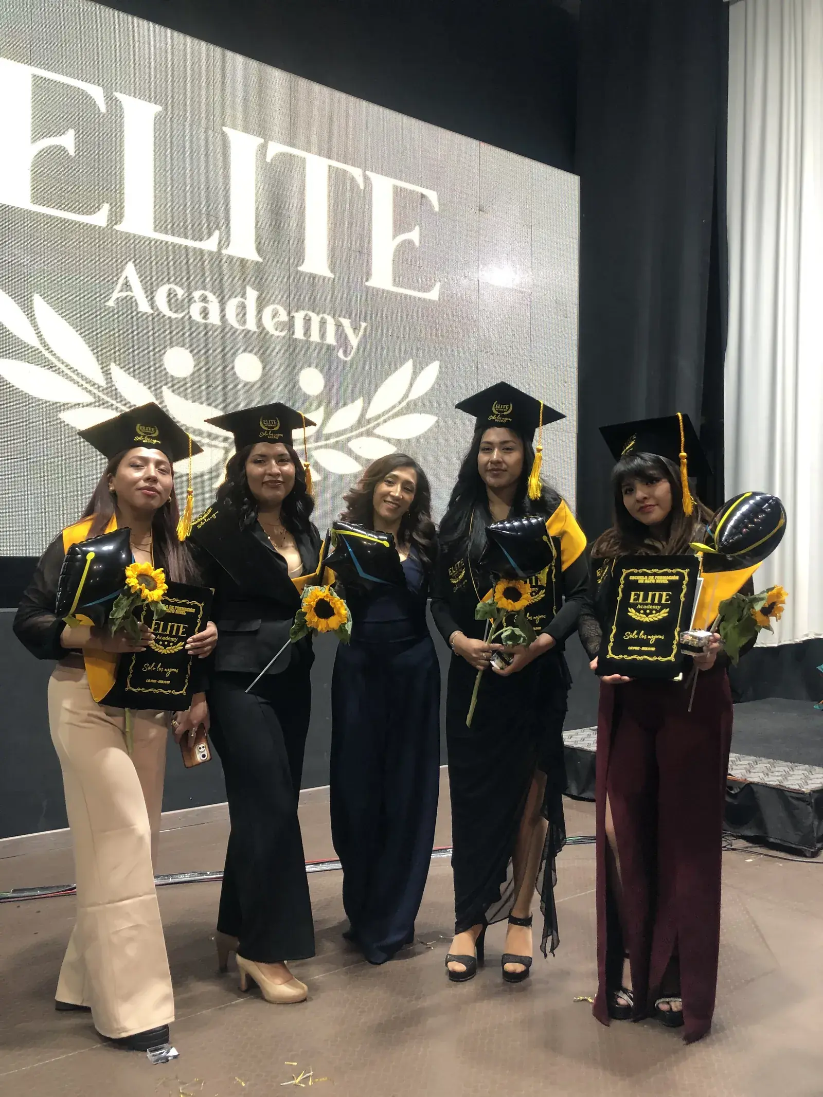 Egresadas con girasoles frente al letrero de Elite Academy